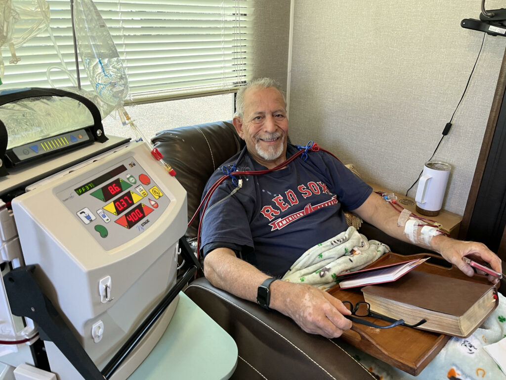 An elderly man is sitting in a recliner, receiving dialysis. He is smiling, wearing a Red Sox T-shirt, and holding a book and glasses on his lap. Medical equipment is beside him, and he is covered with a blanket.
