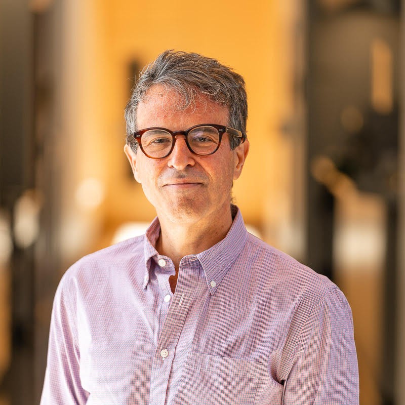 A man with gray hair and glasses, wearing a light purple button-up shirt, stands indoors with a softly blurred, warm-toned background. He is looking at the camera and smiling slightly, representing Renal Associates.