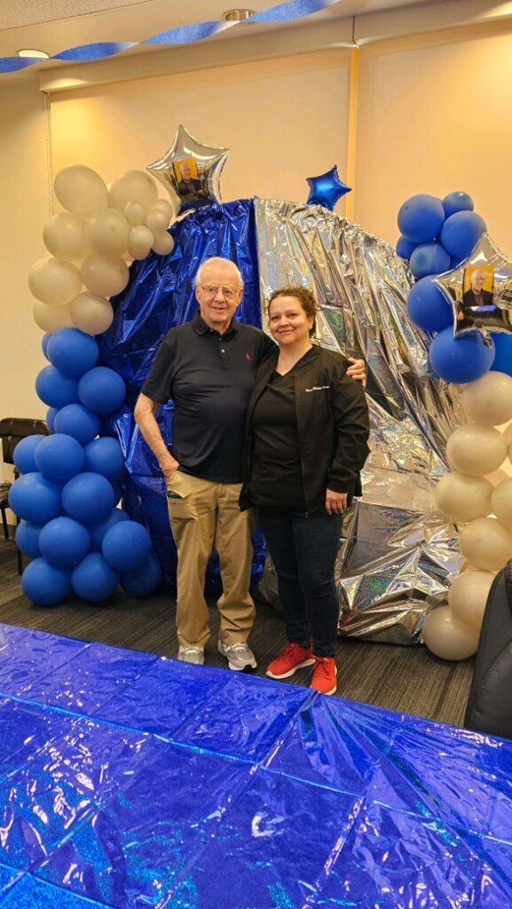 Dr. Dale Erikson and a guest stand smiling in front of a festive retirement backdrop decorated with blue and white balloons, silver stars, and shiny metallic fabric. The man wears a navy shirt and khaki pants; the woman wears a black jacket and red shoes.