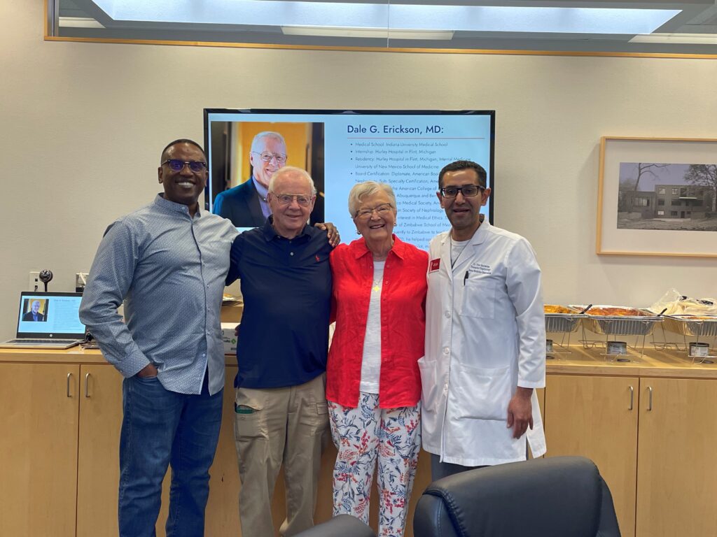 Four people stand together smiling in an office; a presentation slide honoring Dr. Dale Erikson’s retirement is displayed on a screen behind them. Food trays are on a table in the background.