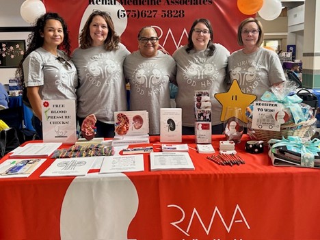 Five women in matching gray shirts stand behind a table with kidney health materials, a raffle basket, and medical displays. The tablecloth and banner read "Renal Medicine Associates." The group smiles at the camera. Five women in matching gray shirts stand behind a table with kidney health materials, a raffle basket, and medical displays. The tablecloth and banner read "Renal Medicine Associates." The group smiles at the camera.