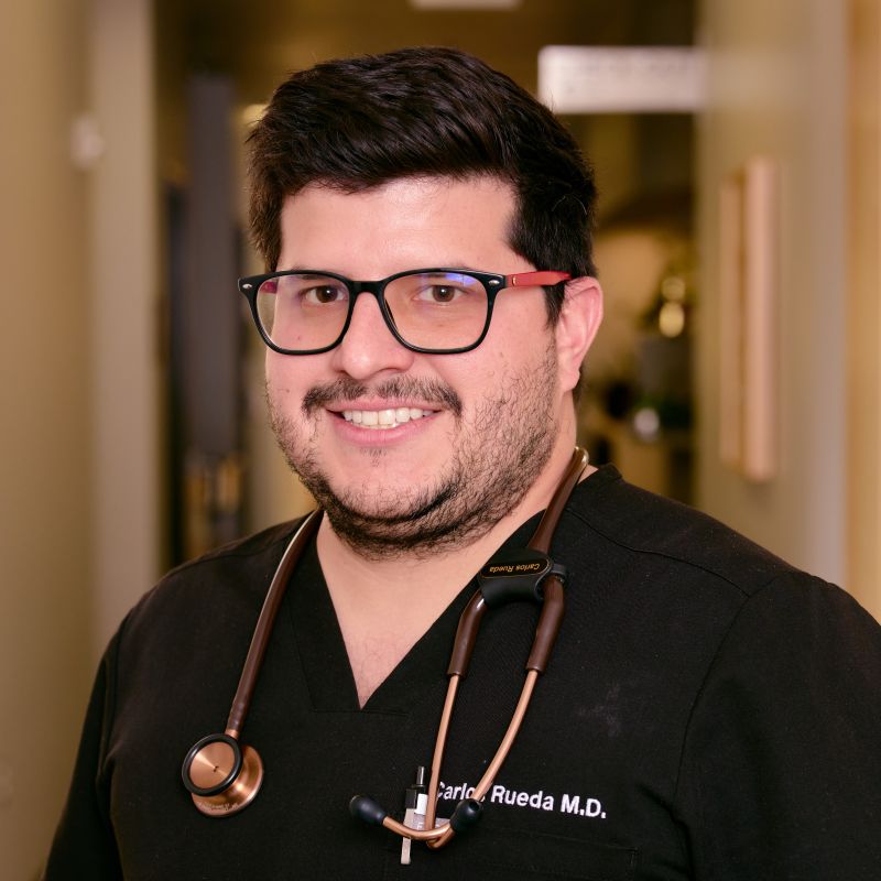 A man with dark hair, a beard, and glasses is smiling. He is wearing black medical scrubs with a stethoscope and the name "Carlos Rueda M.D." embroidered on his shirt. The Renal Associates hallway forms the background.
