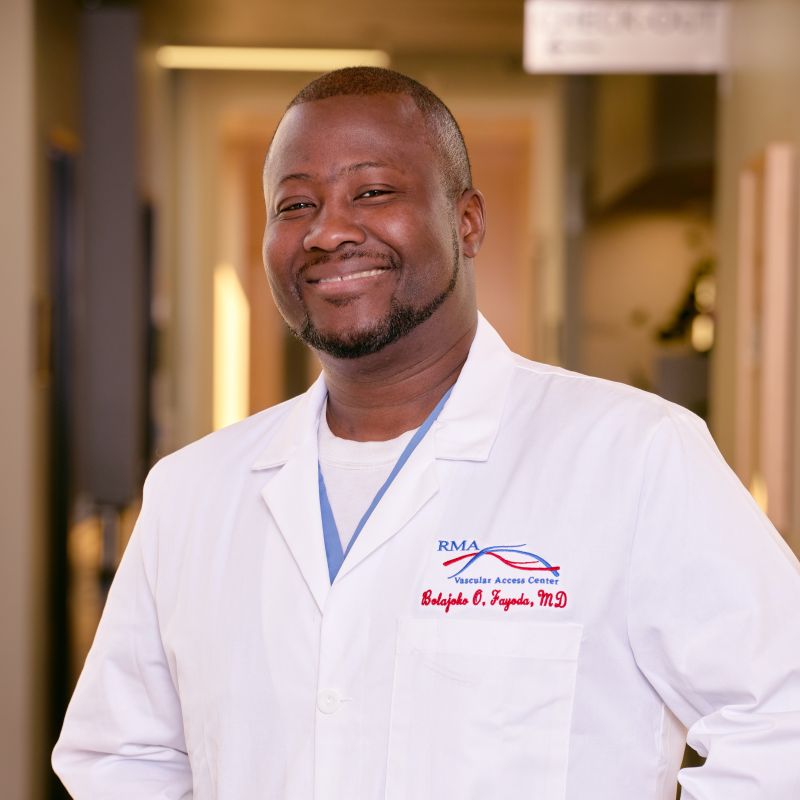 A smiling man wearing a white lab coat with "RMA Vascular Access Center," "Renal Associates," and "Babafola O. Fagade, MD" embroidered in red, standing in a well-lit hallway.