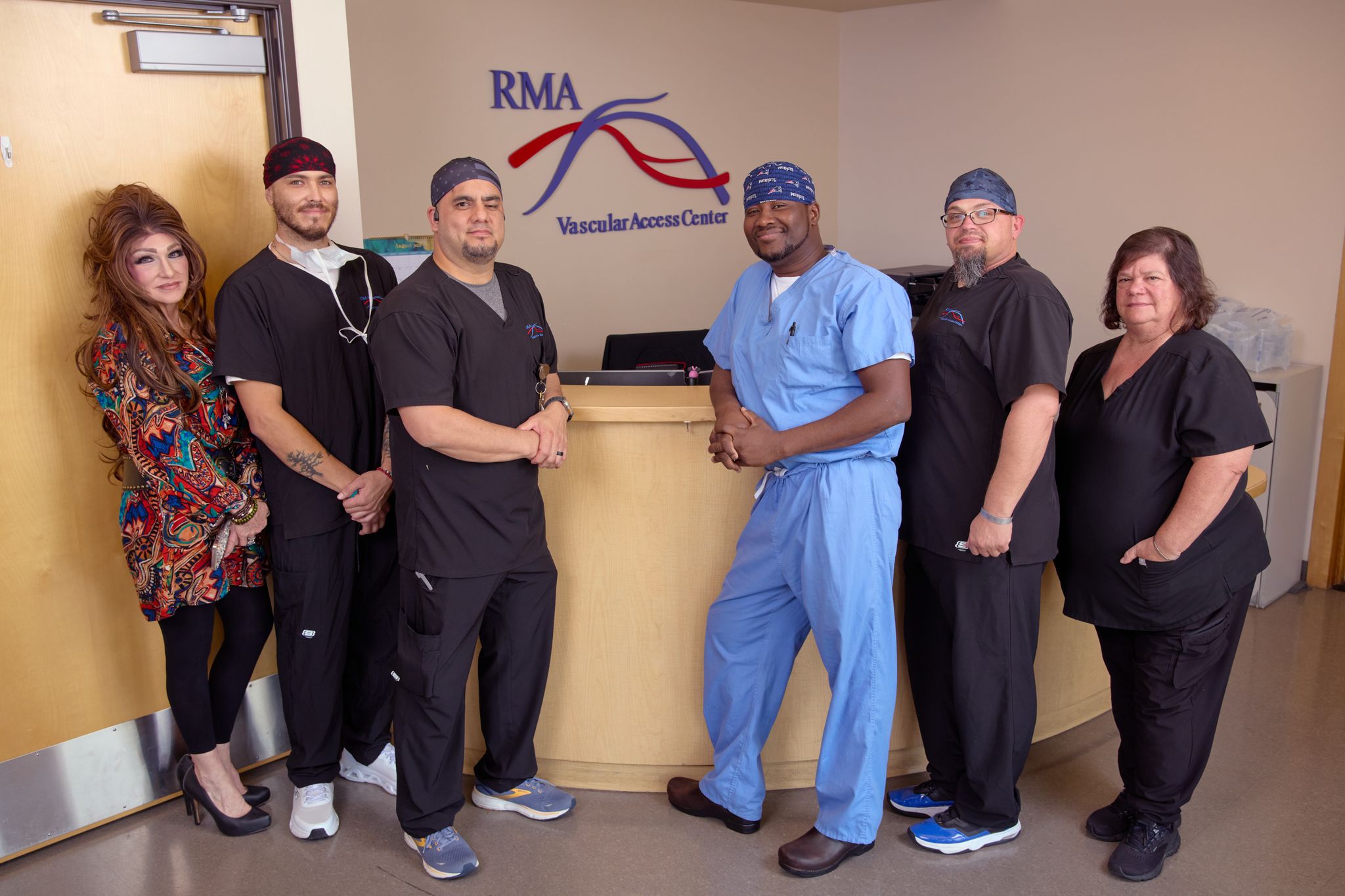 Six medical professionals stand in front of the Access Center reception desk at RMA Vascular Access Center. Five wear black scrubs, one wears blue, and they pose together, smiling, in a clinical setting.