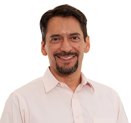 Smiling man with dark hair and a goatee, wearing a light pink button-up shirt, standing against a plain white background.