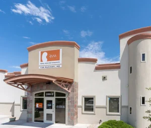 A modern medical office building with a sign reading "RMA Royal Medicine Associates Tim Muchayi, MD" above the entrance, under a clear blue sky with scattered clouds.