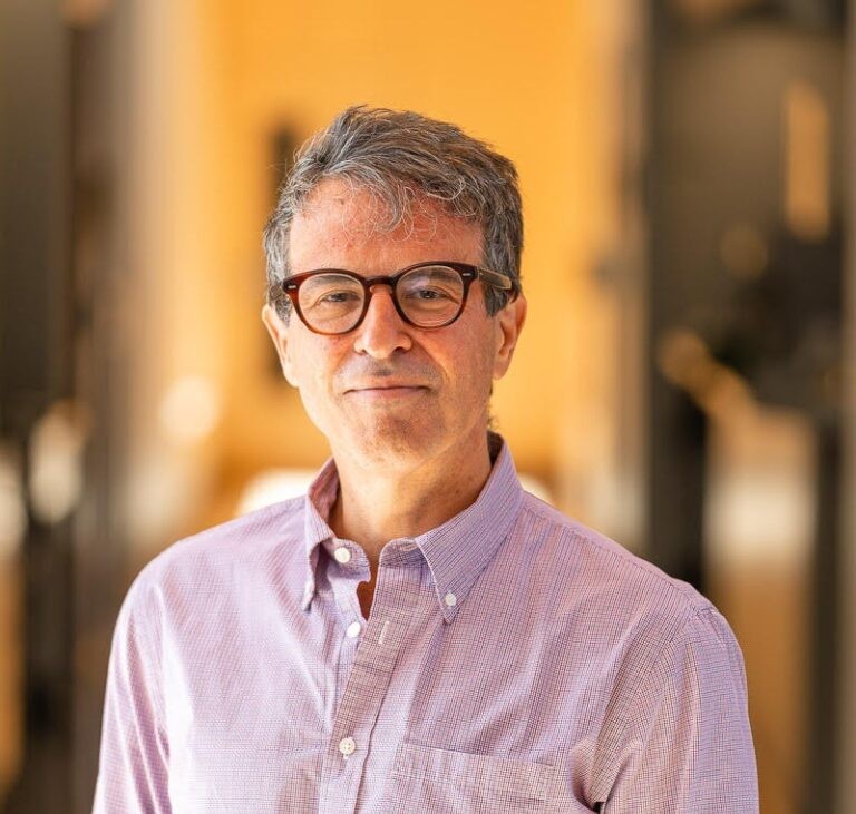 A middle-aged man with short gray hair and glasses, wearing a light purple checkered button-up shirt, stands indoors in Northeast Albuquerque with a softly blurred background in warm tones.
