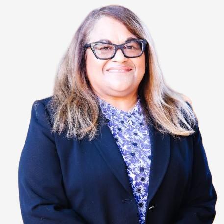 A woman with long, light brown hair and glasses is smiling. She is wearing a black blazer over a blue patterned blouse and is posed against a plain, light background.