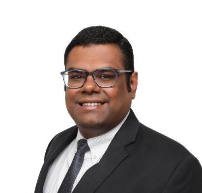 A man from Northeast Albuquerque wearing glasses, a dark suit, white shirt, and polka dot tie, smiling at the camera against a plain white background.