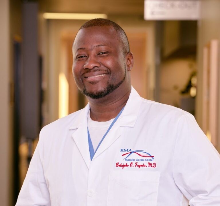 A smiling man wearing a white lab coat stands in a softly blurred hallway in Northeast Albuquerque. The coat is embroidered with “RMA” and “Babajide O. Fagodu, M.D.” Subtle office elements appear in the background.