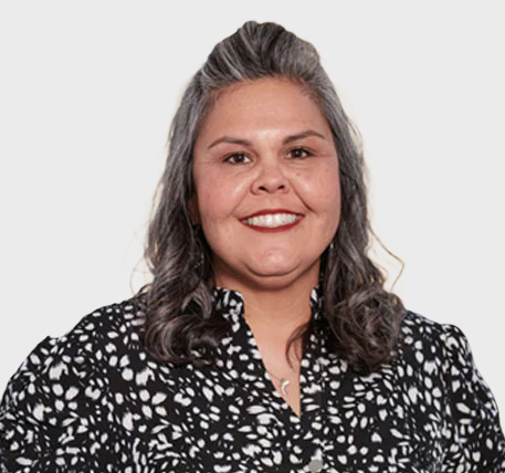 Smiling woman with wavy gray hair wearing a black and white patterned blouse, standing against a plain light background.