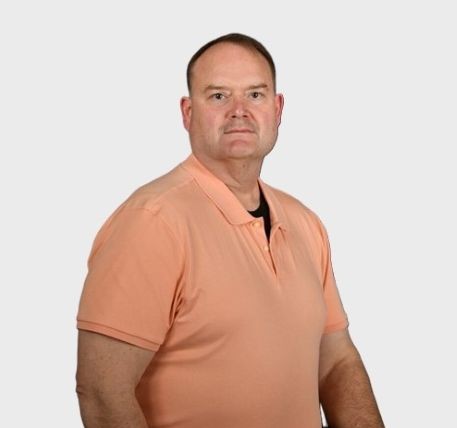 A middle-aged man with short hair, wearing a peach-colored polo shirt, stands against a plain light gray background, looking directly at the camera with a neutral expression.