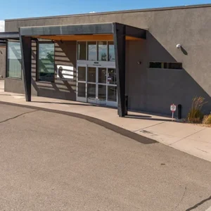 A modern, single-story building in Northeast Albuquerque with gray walls, a flat roof, and a large glass entrance door, surrounded by a paved sidewalk and minimal landscaping.