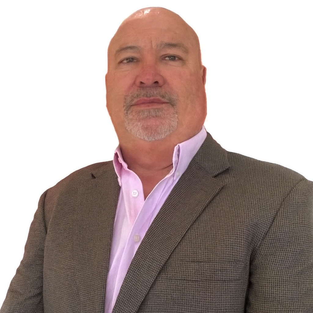 A middle-aged man with a bald head and gray goatee, wearing a gray blazer over a light pink dress shirt, poses against a plain white background.