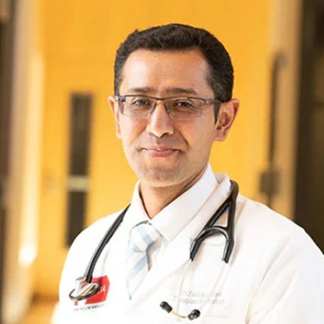 A male doctor with short dark hair and glasses is wearing a white coat, light blue striped tie, and a stethoscope around his neck, standing in a brightly lit hallway.