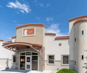 A modern beige and white building with an arched entrance, large windows, and an orange sign featuring a white silhouette of a head and the letters "RMA" under a blue sky with scattered clouds.