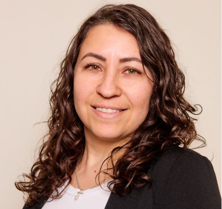 A woman with long, curly brown hair smiles slightly at the camera. She is wearing a black blazer over a white top and a silver necklace. The plain, light background suggests a professional portrait for Advance Care Practitioners in New Mexico.