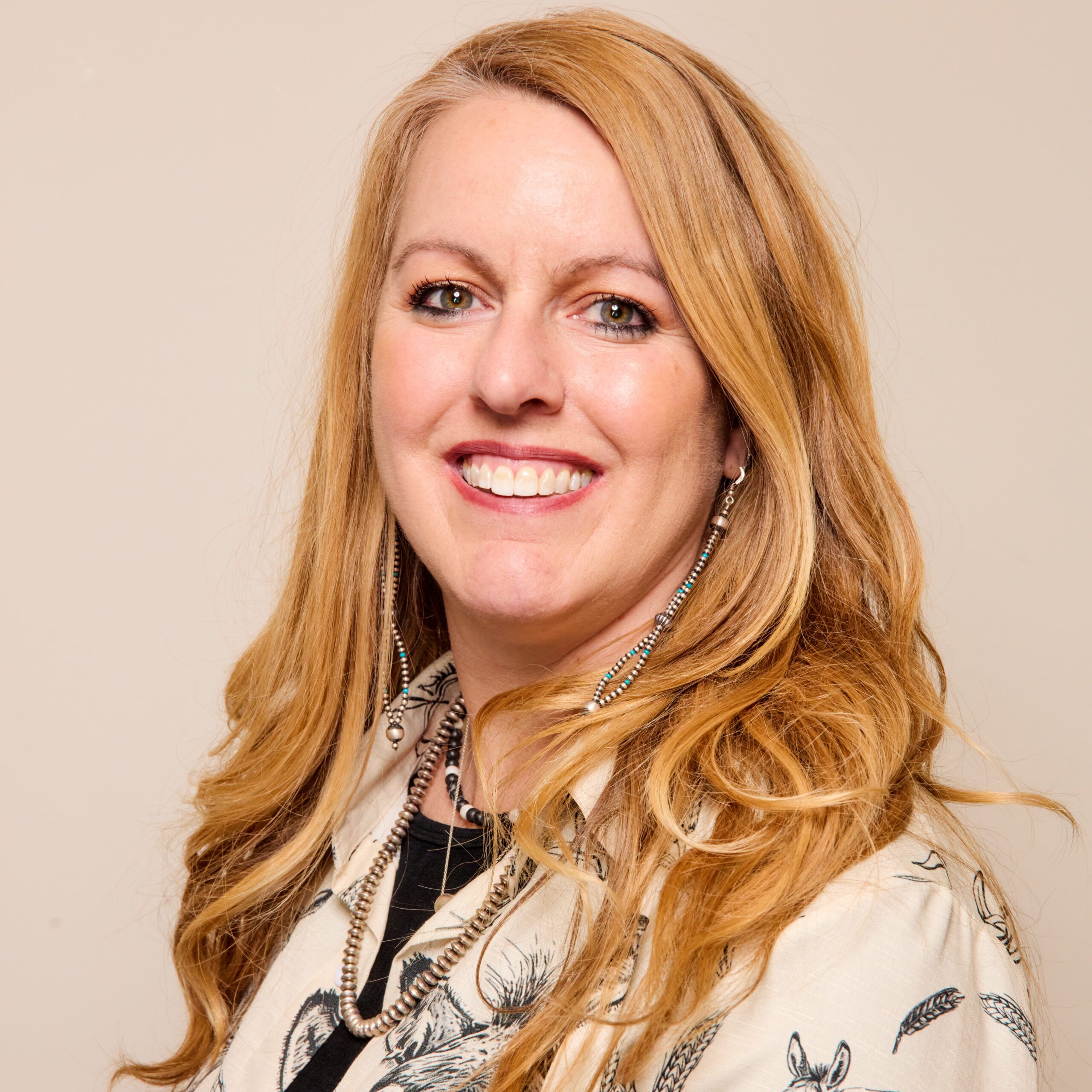 A woman with long, wavy blonde hair smiles at the camera. She is wearing a light-colored top with a black and white pattern and long, dangling earrings. The background is plain and neutral.