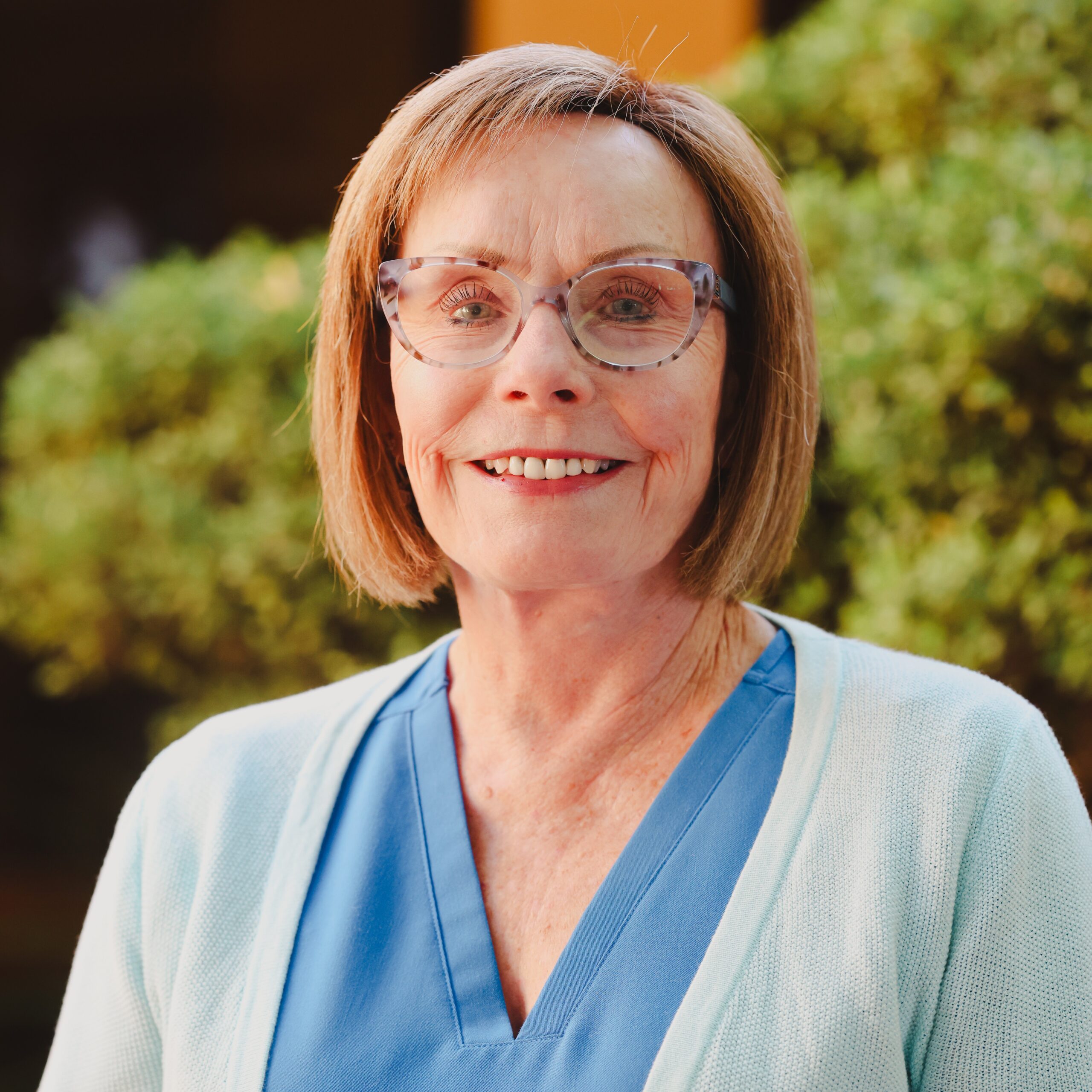 An older woman with short light brown hair, glasses, and a blue V-neck top with a light cardigan smiles outdoors in front of blurred greenery, reflecting the approachable spirit of Advance Care Practitioners & physician assistants in New Mexico.