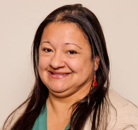 A woman with long dark hair smiles at the camera. She is wearing a beige sweater, a green top, and red dangling earrings, with a plain light background—representative of Advance Care Practitioners & physician assistants in New Mexico.