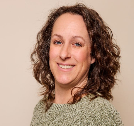 A woman with wavy brown hair and light skin smiles at the camera. She is wearing a green textured sweater and standing against a plain beige background, representing Advance Care Practitioners & physician assistants in New Mexico.