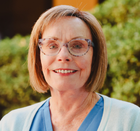 A smiling older woman with short light brown hair and glasses, wearing a blue top and light cardigan, stands outdoors in front of blurred greenery, representing Advance Care Practitioners & physician assistants in New Mexico.
