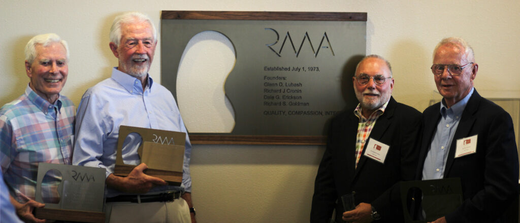 Four older men smile and pose holding awards in front of a wall plaque displaying the RMA logo and founders' names. They are dressed in button-down shirts and blazers, standing indoors.