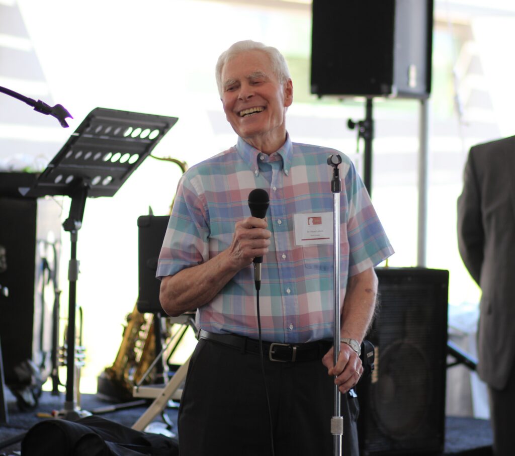An older man in a colorful plaid shirt stands smiling with a microphone in hand, speaking at an event with musical instruments and equipment visible in the background.