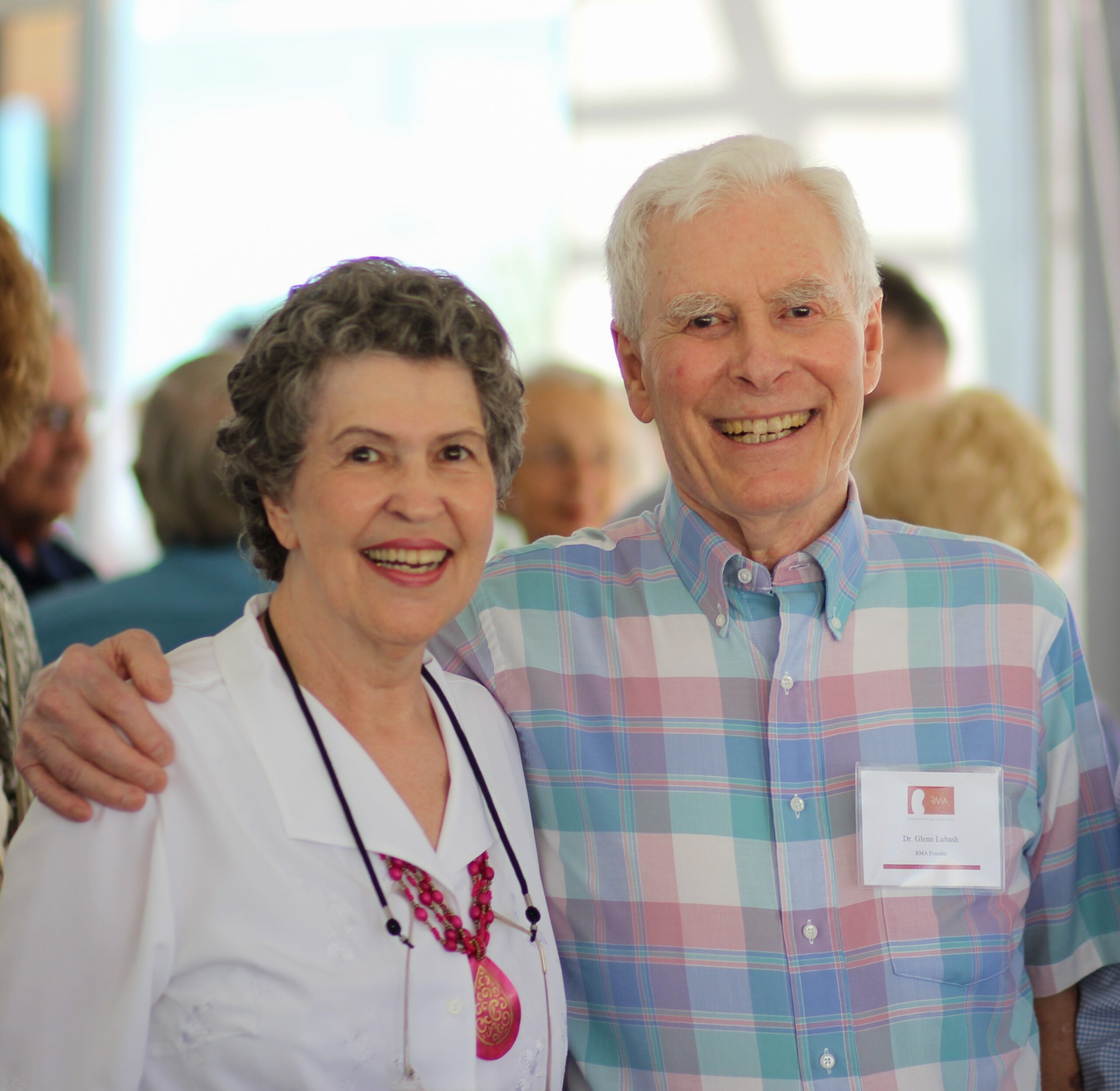 An older woman and man stand smiling together at an event. The man has his arm around the woman. Both are dressed casually, and people are visible in the blurred background.