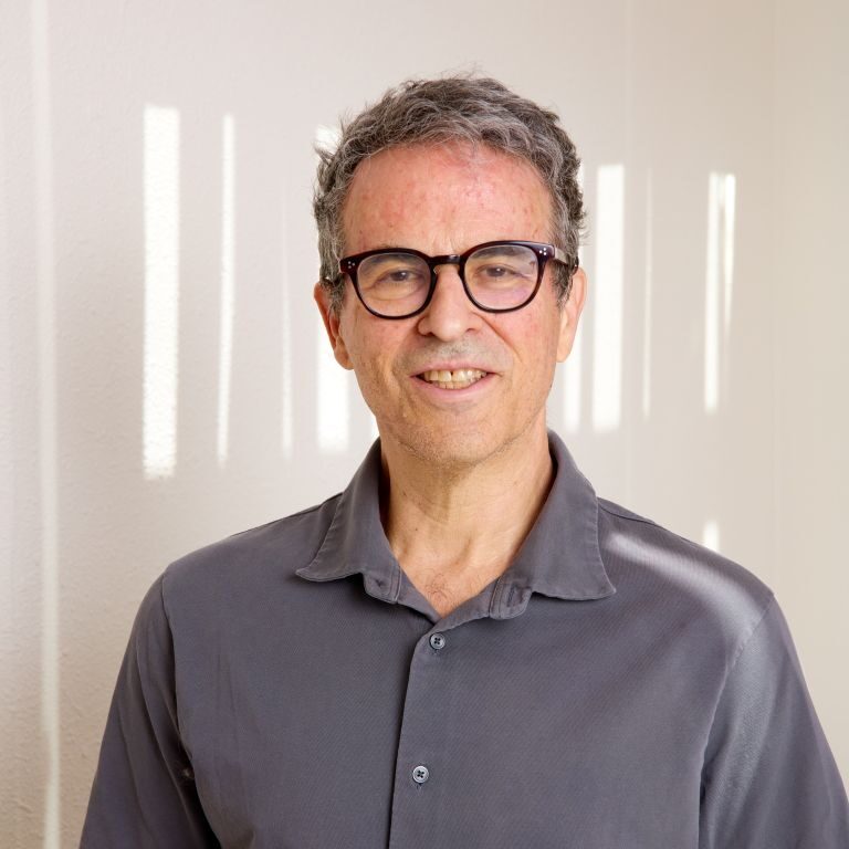 A middle-aged man with short gray hair and glasses smiles while standing indoors, wearing a gray collared shirt. Sunlight filters through blinds, creating striped shadows on the wall behind him—a warm atmosphere that reflects the welcoming environment at Santa Fe Nephrology.