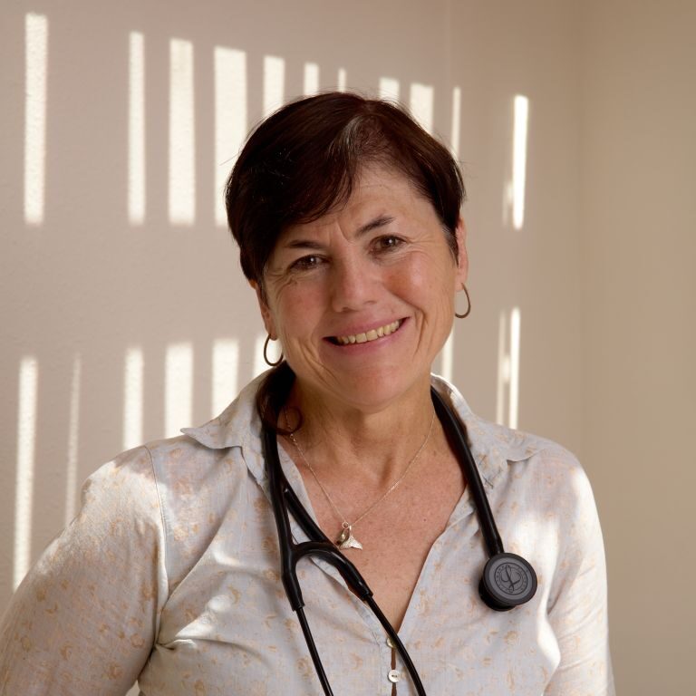 A woman with short dark hair, wearing a light blouse and a stethoscope around her neck, stands smiling in a sunlit room with shadows on the wall, embodying the compassionate care at Santa Fe Nephrology.
