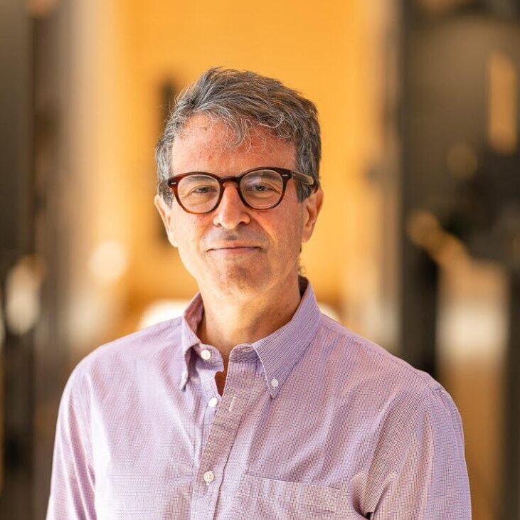 A middle-aged man with short gray hair and glasses, wearing a light purple checkered button-up shirt, stands indoors in Northeast Albuquerque with a softly blurred background in warm tones.