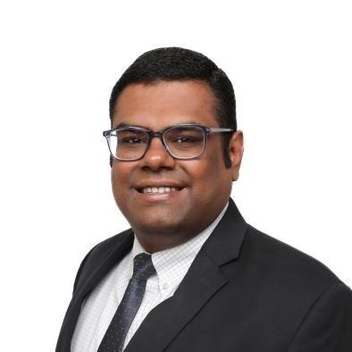 A man from Northeast Albuquerque wearing glasses, a dark suit, white shirt, and polka dot tie, smiling at the camera against a plain white background.