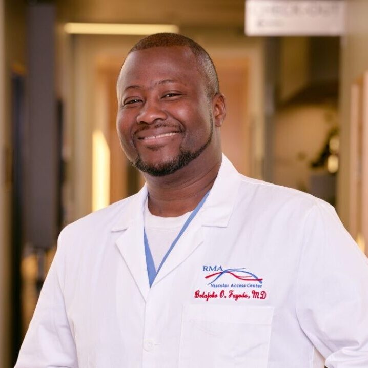 A smiling man wearing a white lab coat stands in a softly blurred hallway in Northeast Albuquerque. The coat is embroidered with “RMA” and “Babajide O. Fagodu, M.D.” Subtle office elements appear in the background.